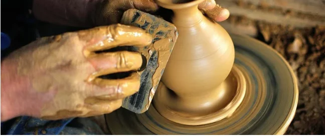 A potter's hands shaping a vessel on the wheel with a metal rib in a Vietnamese craft village